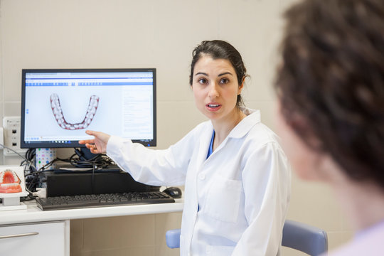 Young female stomatologist in medical gown sitting on chair and showing patient teeth on computer in dentistry