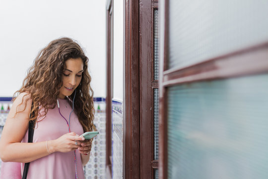 Attractive dreaming brunette with long curly hair listing to music by smartphone standing beside glass doors