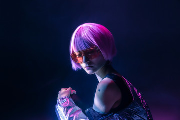 Trendy young Japanese woman with purple hair standing in sparkly silver jacket and red sunglasses sitting on blue background