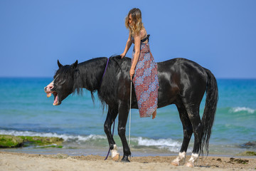 Plakat Portrait of a beautiful blond girl sitting on the back of her black horse on the sea beach