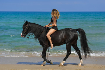 Fototapeta na wymiar Portrait of a beautiful blond girl sitting on the back of her black horse on the sea beach