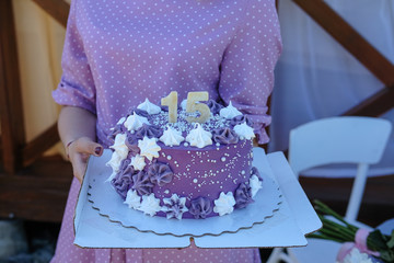 Young beautiful girl in a purple dress holding a birthday cake with the number fifteen, decorated with purple cream and meringue