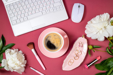 Fashion feminine workspace flat lay. Female home office with laptop, coffee cup, peony flowers, jewelry accessories and cosmetics on burgundy background. Feminine background. Top view