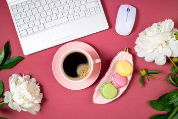 Fashion feminine workspace flat lay. Female home office with laptop, coffee cup, peony flowers, macaroons cake on burgundy background. Femininity background. Top view