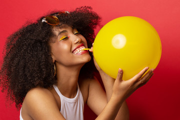 model with a lush hairstyle of curly black hair inflates a yellow balloon on a red Studio background