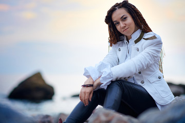 dreadlocks fashionable girl dressed in white jacket and black leather trousers posing near sea in...