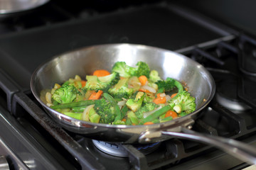 Stir fry vegetables cooking in a stainless steel pan on a natural gas stove.