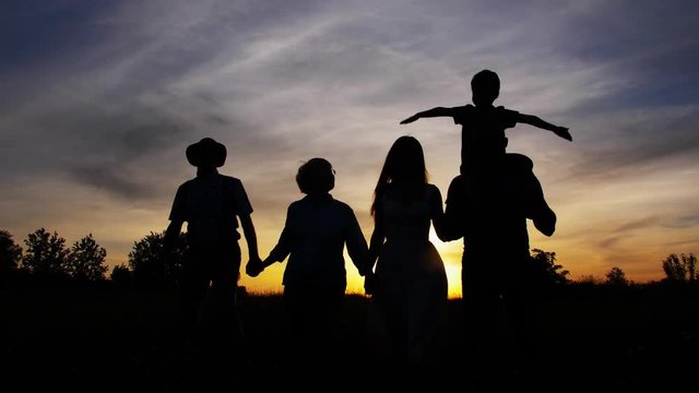 Silhouettes of three generation family enjoying joint countryside leisure. Elderly grandparents, adult mother and father walking hand in hand in meadow at sunset, little son sitting on dad's shoulders