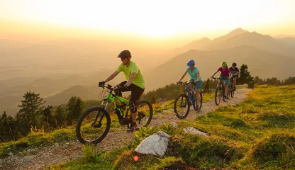Foto op Canvas SUN FLARE Cheerful tourists ride electric bicycles up a mountain trail at sunset © helivideo