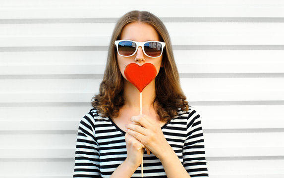 Portrait close-up woman kissing red heart shaped lollipop or hides her lips on white wall background
