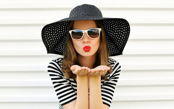 Portrait woman blowing red lips sending sweet air kiss in black summer straw hat on white wall background