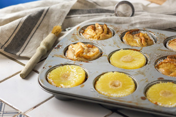 Gluten-free homemade vegetarian desserts cupcakes muffins with apple slices and pineapple rings on top in baking tray in kitchen on white-tiled worktop, next to a kitchen tea-strainer, towel