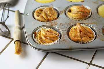 Gluten-free homemade vegetarian desserts cupcakes muffins with apple slices and pineapple rings on top in baking tray in kitchen on white-tiled worktop, next to a kitchen tea-strainer, towel