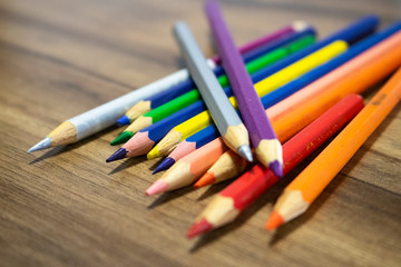 Lápis coloridos dispostos em cima de uma mesa com textura amadeirada. Cores dos lápis azul, roxo, rosa, vermelho, laranja e prata.