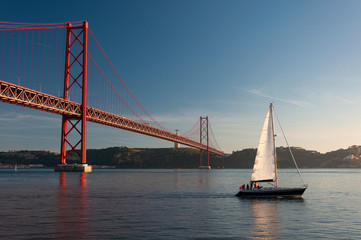 Fototapeta na wymiar Sailing boat passing by the 25 of April Bridge (Ponte 25 de Abril) over the Tagus River in the city of Lisbon, Portugal; Concept for travel in Portugal