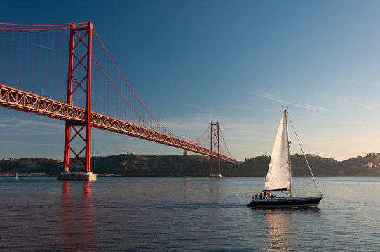Sailing boat passing by the 25 of April Bridge (Ponte 25 de Abril) over the Tagus River in the city of Lisbon, Portugal; Concept for travel in Portugal