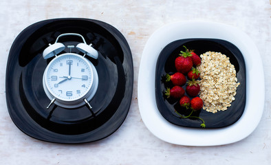 Black plate with oatmeal and strawberries on a white background. Alarm clock shows 8 am. Healthy breakfast time. The concept of healthy eating.