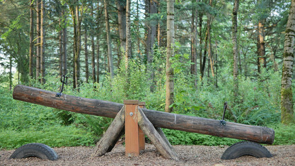 A wood log seesaw teeter totter in a park playground with green forest background.