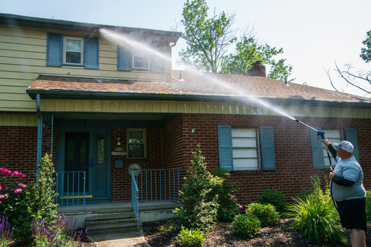 One heavy overweight Caucasian man spraying water solution on the front of a house as part of his pressure washing service. The house has some siding but is mostly red brick. There are blue shutters.