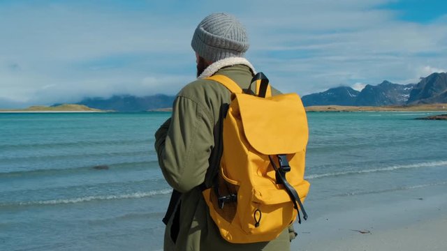 Traveler in a green jacket and a yellow backpack stands on the beach and against the backdrop of high mountain peaks, inspired by the epic landscape of Lofoten Islands, Norway