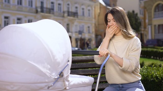 Yawning mother swinging infant carriage sitting on park bench, lack of sleep