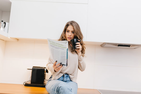 Lovely girl wears jeans expressing amazement while reading newspaper. Indoor photo of elegant curly woman with cup of tea holding magazine in her kitchen.
