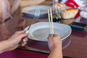 Female hand is holding chopsticks and fork over an empty plate.