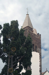 the steeple of funchal cathedral in madeira behind a tree with decorative lights with blue cloudy...