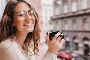 Emotional white woman wears glasses posing on blur background with cup of hot beverage. Charming smiling curly lady spending time near window, looking at city and drinking tea.