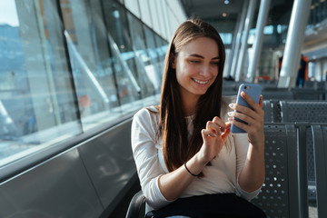 Young beautiful woman sitting at the gate and using smartphone while waiting for a flight at the airport. Travel concept. Passenger girl chatting and smiling in terminal departure lounge.