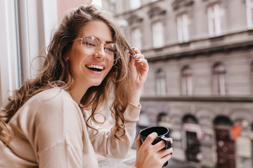 Close-up portrait of laughing brunette girl in beige sweater drinking coffee on city background....