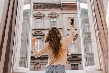 Carefree slim girl in jeans dancing beside window with cup of coffee. Portrait from back of...