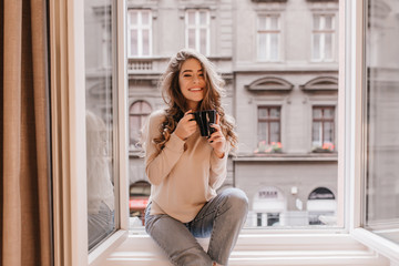 Happy white woman with gorgeous hairstyle sitting on window sill. Indoor portrait of glad young...