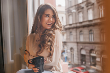 Sensual woman with dark hair looking with excitement at city from window. Indoor portrait of...