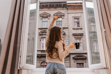Indoor photo from back of trendy woman in jeans drinking tea and looking at city. Inspired female model with curly long hair stretching near window.