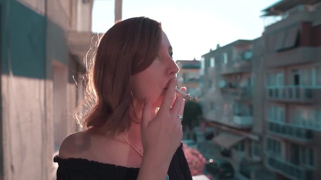 Attractive young girl is smoking on balcony with magnificent sun flare. Summer time, cigarette or weed beauty slow motion