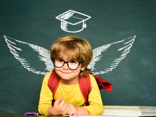 School wings and dream. Cheerful smiling child at the blackboard. Cute boy with happy face expression near desk with school supplies. Blackboard background.