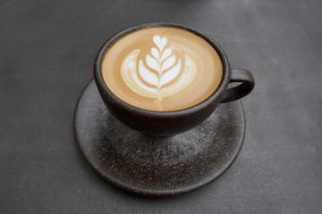 Cappuccino with artful milk foam pattern in a rustic brown cup with saucer. Close-up in front of dark background