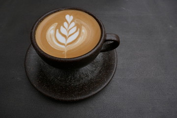 Cappuccino with artful milk foam pattern in a rustic brown cup with saucer. Close-up in front of dark background
