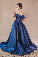Gorgeous brunette curly hair model posing in elegant blue evening dress on the beige paper studio...