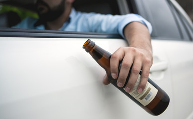 Drunk young man driving a car on the road holding a bottle of beer; Dangerous drunk driving