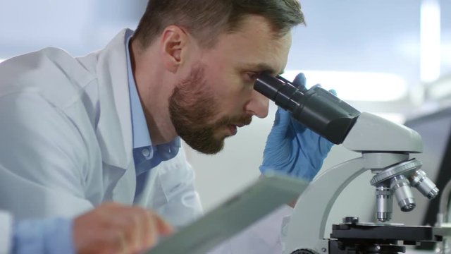 Close up of male and female biochemists in medical gowns standing at desktop in lab. Man working on microscope while his female laboratory assistant writing down results using tablet