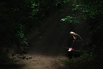 Lonely and sad blonde woman sitting on a bench in a light spot in the middle of dark forest path, surrounded with green trees