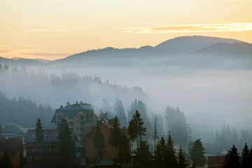 Afwasbaar Fotobehang Mistig bos Resort village houses buildings on background of foggy blue mountain hills covered with dense misty spruce forest under bright pink sky at sunrise. Mountain landscape at dawn.