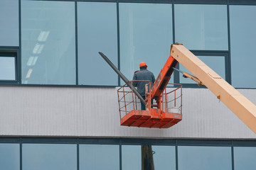 Worker in helmet in aerial platform installing metal sill on external PVC window frame on height. Window sill installation process. Builder in cherry picker inspecting windows