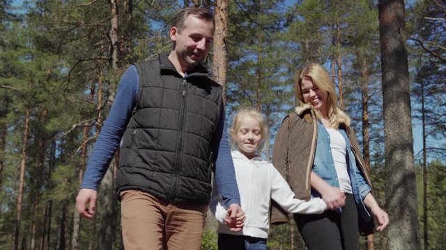 Beautiful happy family of mother, father and daughter walking among pine trees in countryside holding hands. Parents and girl talking cheerfully and smiling