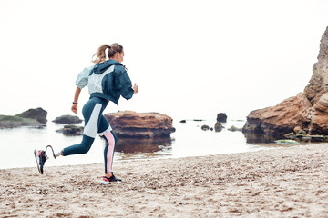 Take care of your body. Side view of strong disabled woman in sportswear with prosthetic leg is running on the beach.