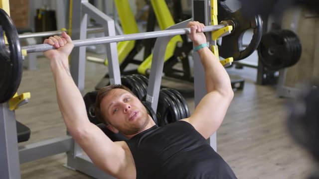 Rack focused shot of professional bodybuilder explaining and showing lying barbell bench press in gym while recording video lesson with professional camera