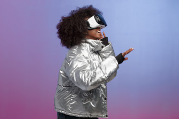 Trendy curly brown-haired girl dressed in a silver-colored jacket uses the virtual reality glasses in the studio on neon background