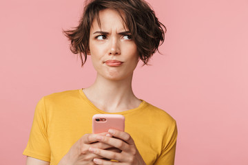 Confused young beautiful woman posing isolated over pink wall background using mobile phone.